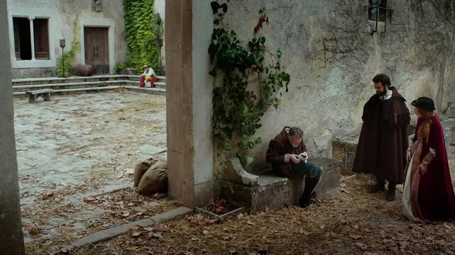 Two characters interact in a small courtyard as one gestures towards the ground while another listens intently. The location shows old stone buildings and scattered leaves.
