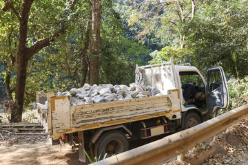 Rugged white truck hauls heavy cargo of stone on rural dirt road through dense forest. Transportation work showing construction and industry in remote area