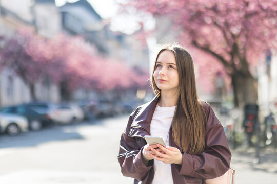 Young woman with smartphone on spring city streets. Girl walking, using map app, enjoying cherry blossoms. Urban travel, slow living lifestyle, mindfulness in springtime.