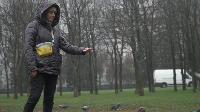 A girl in a black hooded jacket and jeans feeds pigeons with her outstretched arm in a city park in autumn. The birds in flight approach the hand, but are afraid.