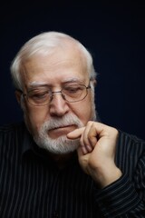 Obraz premium Elderly man sitting quietly with hand on chin, lost in thought, in a dark room during the evening, showing deep contemplation