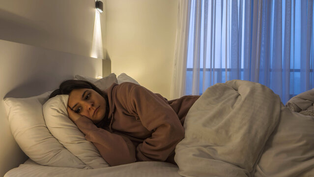 Young woman lying on bed at night with sad expression low mood dim bedroom light emotional exhaustion mental health struggle isolation loneliness depression concept fatigue insomnia