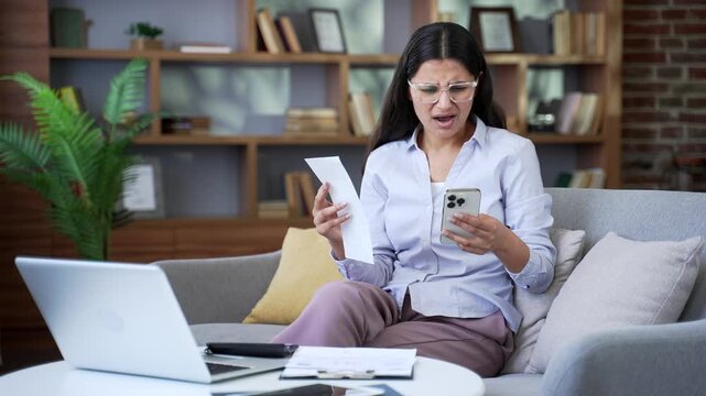 Confused adult woman having difficulty doing household finances using an application on mobile phone sitting on sofa in living room at home office. Shocked female looks at large utility bills.