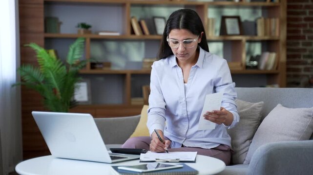 Young woman dealing with household finances using website or app on laptop sitting in home office. Confident adult female holds utility bill and forms payments online by entering data on computer.