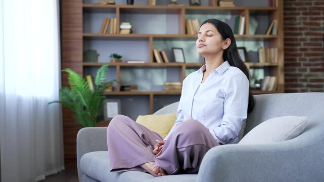 Smiling woman meditating in lotus position sitting on sofa in living room at home. Calm young female practices yoga with closed eyes, breathes deeply, relaxes in cozy atmosphere on comfortable couch.