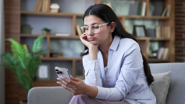Tired bored young woman using smartphone while sitting on sofa at home. Exhausted sad female browsing social media, scrolling feed reading posts, wasting time on mobile phone in living room. Close up