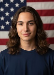 Young adult male with long brown hair smiling in front of a slightly blurred American flag background