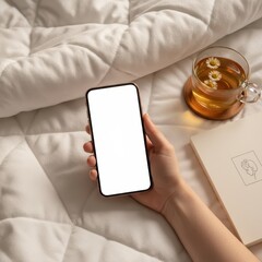 Hand holding black smartphone with blank white screen on quilted white duvet or comforter, with glass cup of chamomile tea and beige book, for app or website mockup