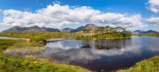 Obraz premium Panoramic mountain lake with Pine Island in Connemara, Galway, Ireland. Calm reflective water under dramatic clouds. Distant rocky peaks, grassy marsh foreground and scattered peat hummocks