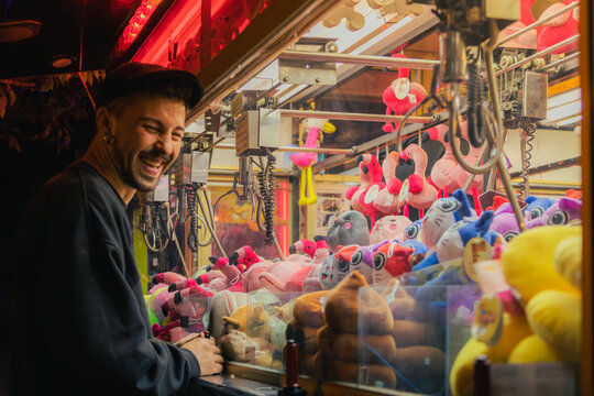 Man smiling in front of a claw machine filled with plush toys. Surrounded by vibrant colored lights.