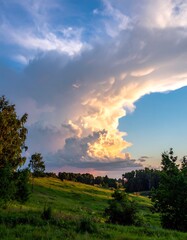 Dramatic sky dominates a lush green landscape. Hues of sunset illuminate fluffy clouds over a rolling hill