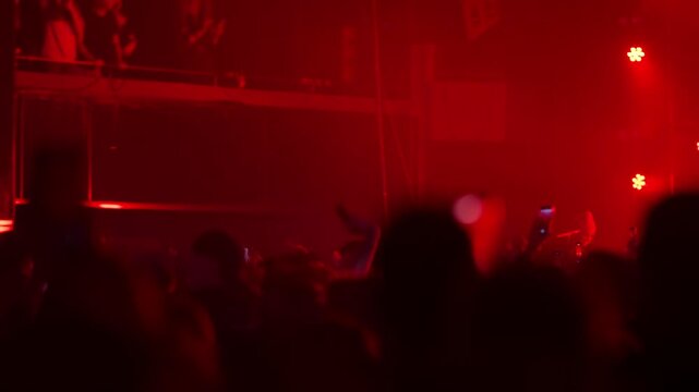 Silhouetted crowd at a live music concert. Fans dance, wave hands, and record with phones as bright blue and red strobe lights flash from the stage, creating an energetic party atmosphere.