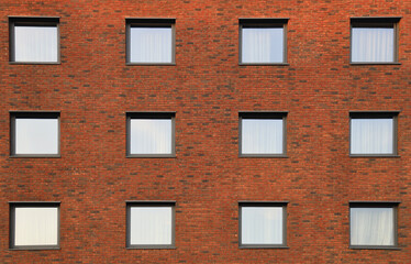 brick fa&ccedil;ade of a multi-story building features numerous windows of same shapes. The warm reddish-orange hue of the brickwork contrasts with the dark window frames.  strict geometric rhythm.