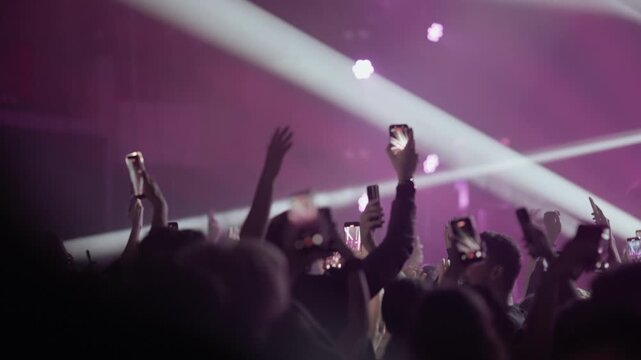 Video of a large crowd at a music festival or rock concert. Fans hold up smartphones to record the band on stage amid dramatic beams of light and smoke. A modern live event experience.