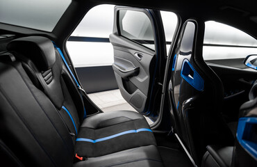 Rear row of seats in a car. Sporty wagon interior with black bucket seats and blue trim elements. Opened rear door is letting bright light from illuminated wall in.
