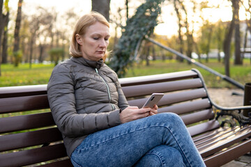 Obraz premium Woman sitting on bench using mobile phone, browsing social media or texting, in autumn park. Remote work and online technology concept