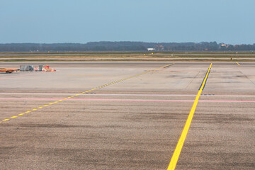 Expansive airport tarmac or taxiway, dominated by painted yellow lines that guide the eye into the distance. The horizon is marked by a line of trees and a flat landscape under a pale sky