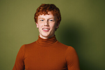 Obraz premium Young redhead man with curly hair wearing orange turtleneck smiling and looking at camera with friendly expression on green background. Portrait of confident male model indoors.
