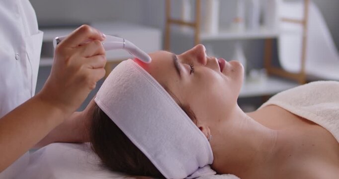 Woman receiving professional red LED light facial therapy, beauty treatment in spa clinic for glowing skin and rejuvenation. Cosmetologist hands using phototherapy device on face skin of lying client