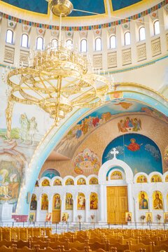 A breathtaking Orthodox church interior showcasing a grand golden chandelier, richly painted biblical frescoes, and an elegant white iconostasis filled with religious icons