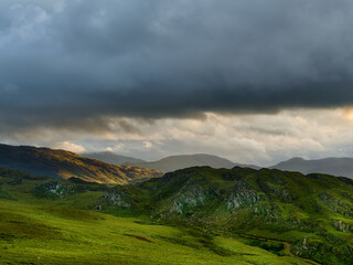 Fototapeta premium Rolling hills under a dramatic, cloudy sky. Green valleys and distant mountain peaks.