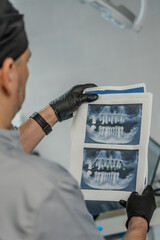 A dentist wearing gloves and a cap holds X-rays of the jaw with the implants marked. A detailed analysis is performed in the office.