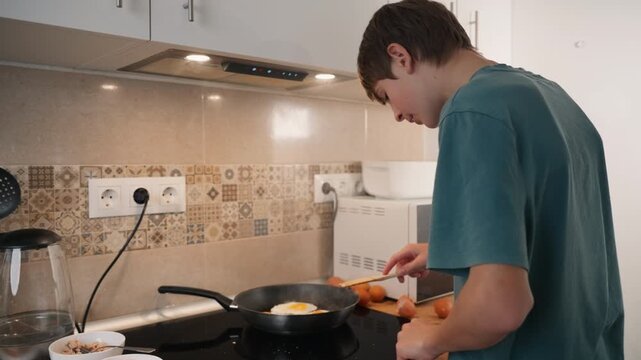 Independent teenager boy cook sunny side up eggs for nutritious breakfast, use spatula in hot pan on modern electric stove, developing essential life and culinary skills