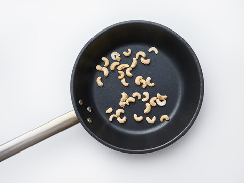 A black frying pan holds several cashew nuts on a white surface. The nuts are scattered and appear uncooked. The setting is bright and simple