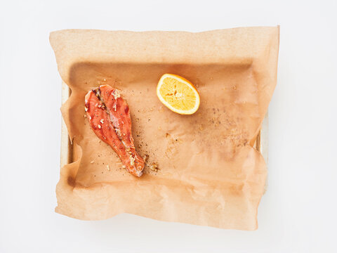 A salmon fillet lies on a tray with parchment paper. A sliced lemon sits nearby. Some spices are scattered on the paper. This scene shows a simple meal preparation