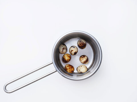 Quail eggs are placed in a metal pot filled with water. The pot is on a white surface, and the scene is captured during daylight. The eggs have variations in color and pattern