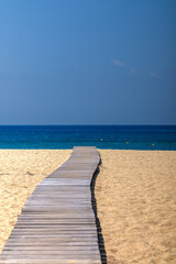 View of a wooden pathway stretching across soft sand towards the open beach of Mylopotas in Ios Greece
