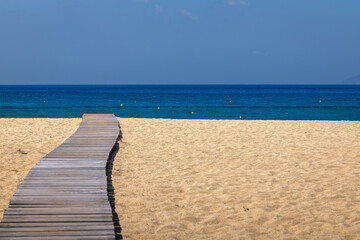 View of a wooden pathway stretching across soft sand towards the open beach of Mylopotas in Ios Greece