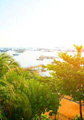 Fototapeta premium View on marina on bright sun. Boats at pier are visible in distance. Fishing boats in sea in bright sunset day. People enjoying view ocean shore. Trees provide green foliage in foreground. Boats dock.