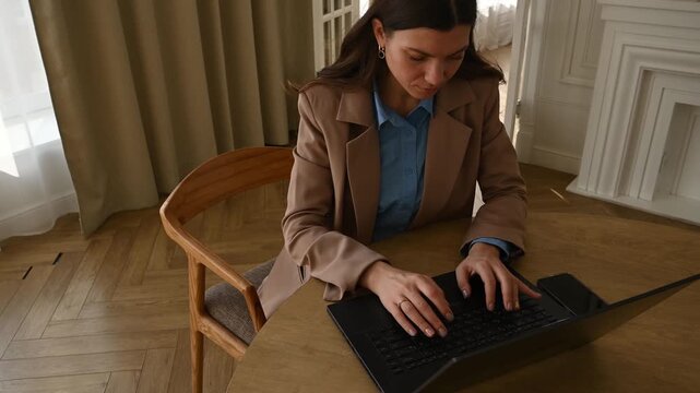 Confident young woman working on her laptop at a wooden table in a bright, modern office, focused on typing an email or report, representing a professional and productive work environment