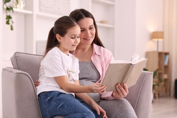 Fototapeta premium Pregnant woman and her daughter reading book together on armchair at home