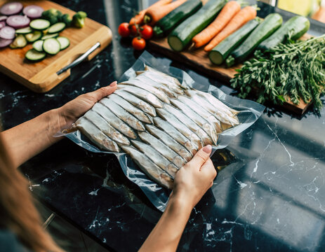 Close-up of female hands holding vacuum-sealed frozen fish on a black marble countertop with fresh vegetables nearby.