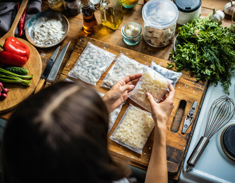Top view of a woman preparing vacuum-packed frozen food portions in a kitchen filled with fresh herbs, oils, and cooking ingredients.