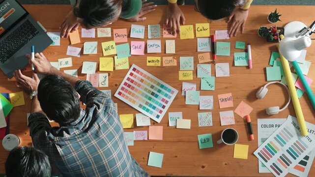 Top down view of business team with casual outfit looking laptop screen and sharing idea. Aerial view of manager working and checking color palette at table with sticky notes and headphone. Symposium.