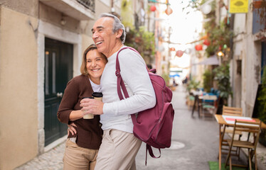 Happy European Senior Couple Traveling Standing And Hugging On Lisbon Street Outside, Holding Paper...