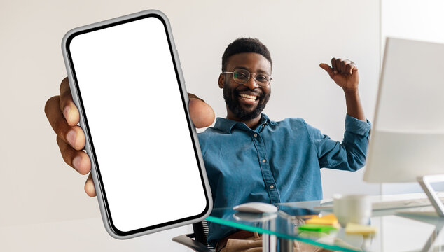 African American man sitting at workdesk with pc, holding a cell phone in his hand, showing blank white screen and gesturing