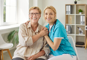 Obraz premium Smiling elderly woman sitting beside cheerful female caregiver in medical scrubs, holding hands with warmth in modern healthcare office. Concept of senior support, trust and professional care 