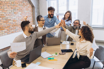 Obraz na płótnie Canvas Diverse business team celebrating success in office, high fiving and cheering during a productive meeting