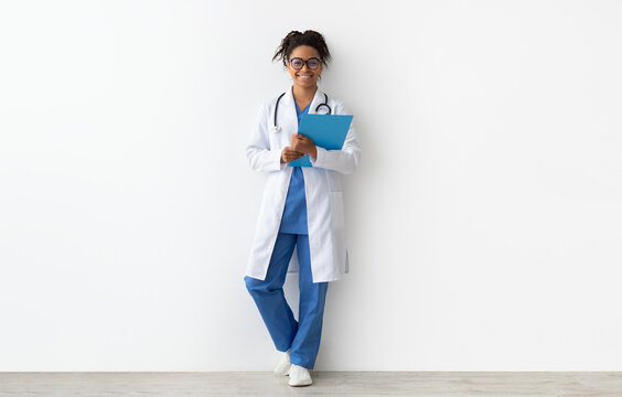 Friendly black doctor in glasses holding clipboard looking at camera standing leaning on wall, lady wearing blue uniform, coat and stethoscope, full body length, white studio background