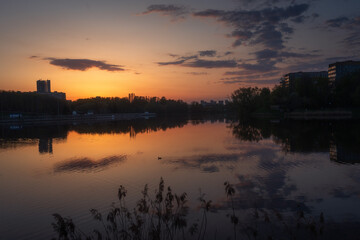 Fototapeta premium An evening city landscape of Minsk at sunset, reflected in a lakeside. Modern buildings and lights shimmer under colorful sky.