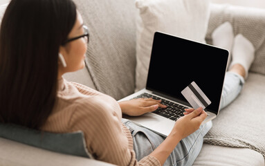 Asian woman making online purchase on laptop computer, holding credit card and listening music