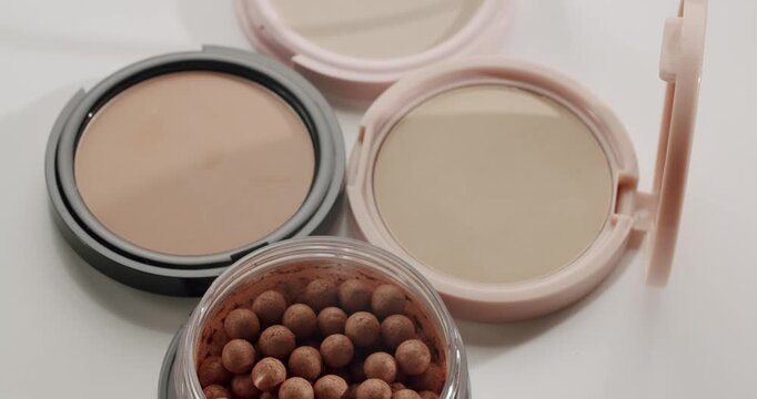 Various professional cosmetic products for face makeup, including pressed powder compacts and shimmering bronzer pearls, are displayed on a clean white background in a beauty studio setting
