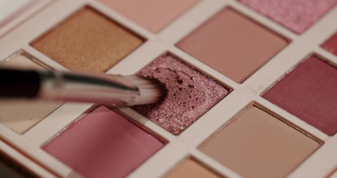 Close-up slow motion shot of a professional makeup artist's brush gathering sparkling pink and rose gold eyeshadow from a colorful palette, showing the texture and pigment of the cosmetic