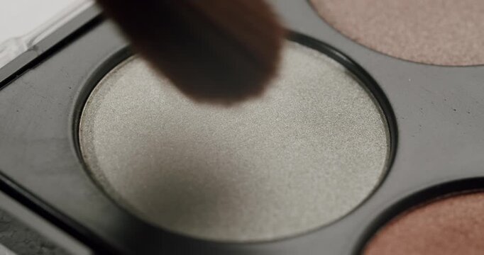 Extreme close-up of a professional makeup brush picking up silver shimmer eyeshadow from a cosmetic palette, demonstrating a beauty routine and application of decorative cosmetics