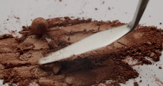Metallic spatula smearing and crushing a brown shimmering cosmetic eyeshadow powder on a white surface, revealing its rich texture and pigmentation in a satisfying macro slow-motion shot