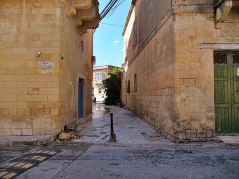 Sunny, daytime scene of Triq San Mikiel street in the Southern Region town
of Zurrieq, Malta. The narrow alley features traditional warm limestone buildings, blue and
green doors, power lines overhead